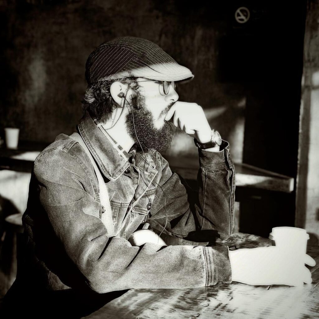 In a quaint café, a bearded man sits at a table, engrossed in his thoughts and savoring a cup of coffee. The atmosphere is captured in a black and white photo, adding a touch of timeless elegance. The man exudes an air of intellectualism as he wears glasses and a hat, his beard giving him a rugged look. His focus is solely on his coffee, which is contained in a white cup that contrasts against the dark table. The café enforces a no smoking policy, as indicated by a sign on a black background. The scene is further enhanced by the presence of a woman, who is also holding a cup of coffee, perhaps engaged in deep conversation with the man. Another black and white photo features a hand holding a cup, creating a sense of poetic contemplation. Overall, the image portrays a contemplative and sophisticated ambiance, inviting visitors to join in the experience.